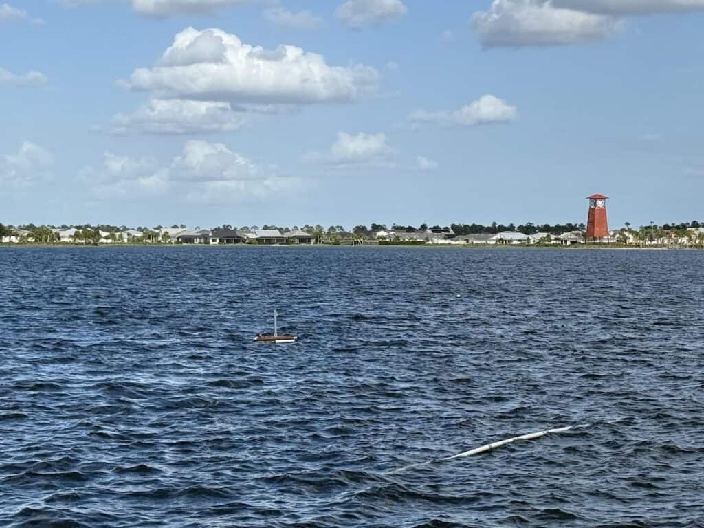 A body of water with a small buoy and a distant shoreline. A red tower and several buildings are visible under a partly cloudy sky.