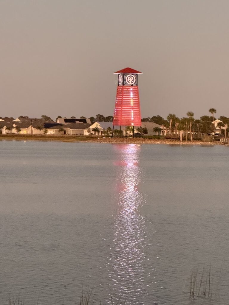 A tall, red cylindrical building with a red roof is illuminated at night by a body of water at Babcock Ranch, with houses and palm trees in the background, embodying the charm often embraced by local small businesses.
