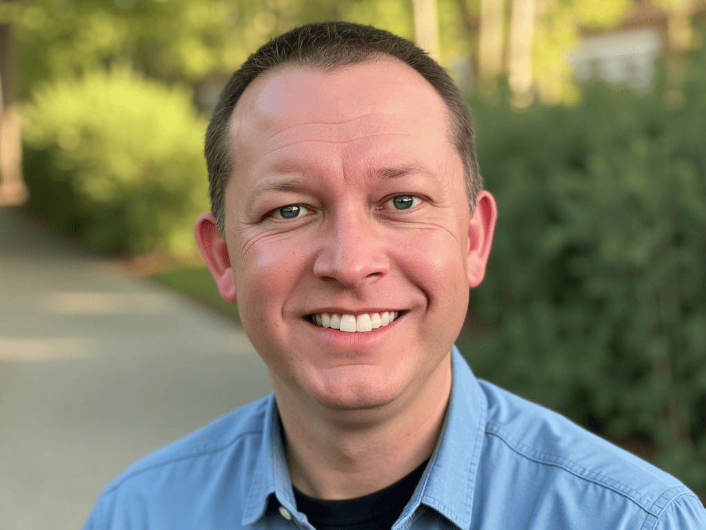 Smiling man in a blue shirt stands outdoors on a sunny day with greenery in the background.